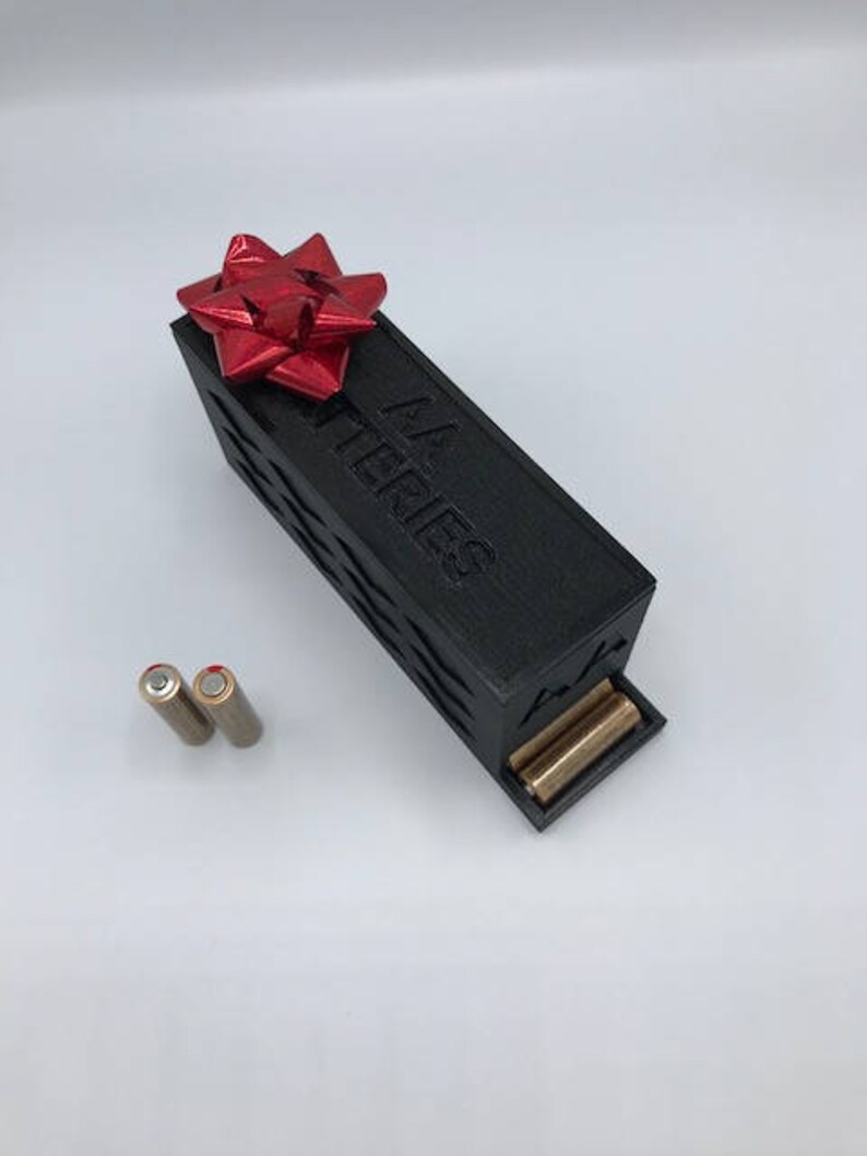 Puede incluir: Una caja negra con un lazo rojo en la parte superior. La caja dice "AA Batteries" en el lateral. Dos pilas AA est&aacute;n al lado de la caja.