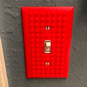 May include: A red light switch cover with a Lego brick pattern. The switch is white and is in the off position.