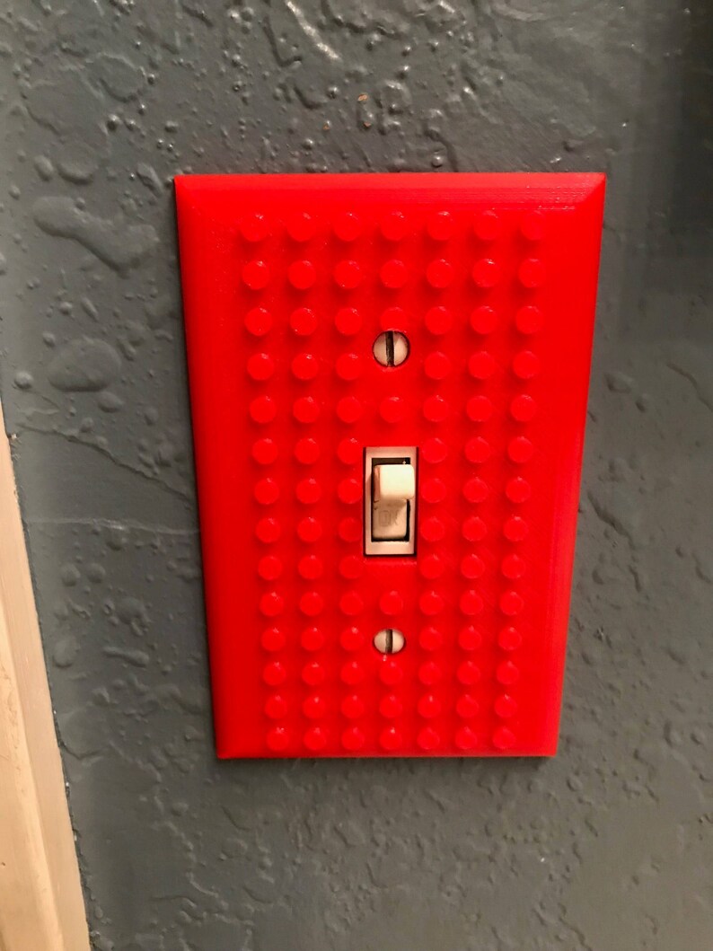 May include: A red light switch cover with a Lego brick pattern. The switch is white and is in the off position.