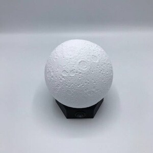 May include: A white moon-shaped night light with a textured surface, sitting on a black base.