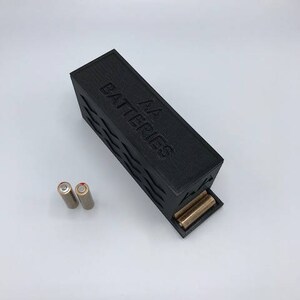 Puede incluir: Una caja de almacenamiento de bater&iacute;as de pl&aacute;stico negro con el texto "AA Batteries" en relieve en la parte superior. Dos bater&iacute;as AA de color dorado son visibles en la caja.