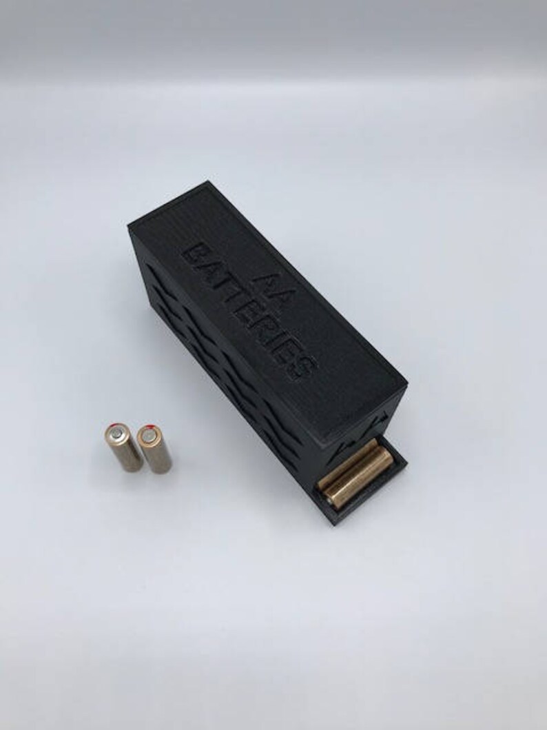 Puede incluir: Una caja de almacenamiento de bater&iacute;as de pl&aacute;stico negro con el texto "AA Batteries" en relieve en la parte superior. Dos bater&iacute;as AA de color dorado son visibles en la caja.