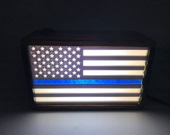 May include: A wooden framed light-up American flag with a blue stripe across the middle, representing the thin blue line.
