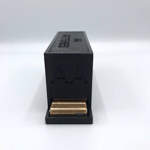 Puede incluir: Una caja de almacenamiento de bater&iacute;as negra con el texto "AA Batteries" en la parte superior y un caj&oacute;n que contiene bater&iacute;as AA.