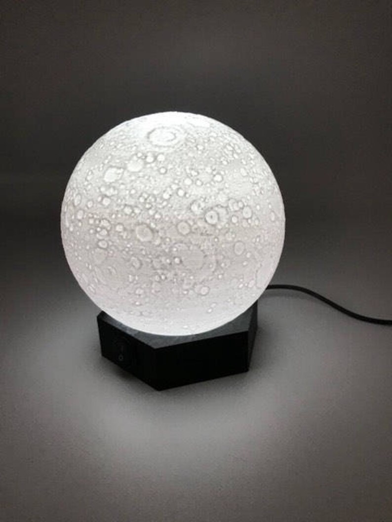 May include: A white moon-shaped lamp with a textured surface sits on a black hexagonal base. The lamp is plugged in and lit, casting a soft glow.