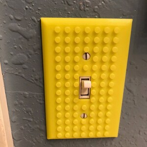 May include: A yellow light switch cover with a Lego-like design. The cover has a white light switch in the center.