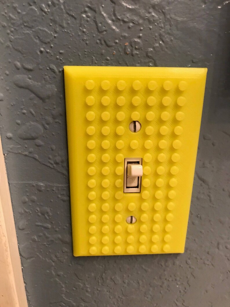 May include: A yellow light switch cover with a Lego-like design. The cover has a white light switch in the center.