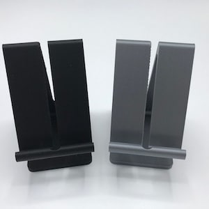 May include: Two phone stands, one black and one gray, with a simple design. Each stand has a horizontal bar across the front to hold a phone.