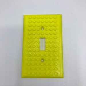 May include: A yellow light switch cover with a textured surface that resembles Lego bricks.