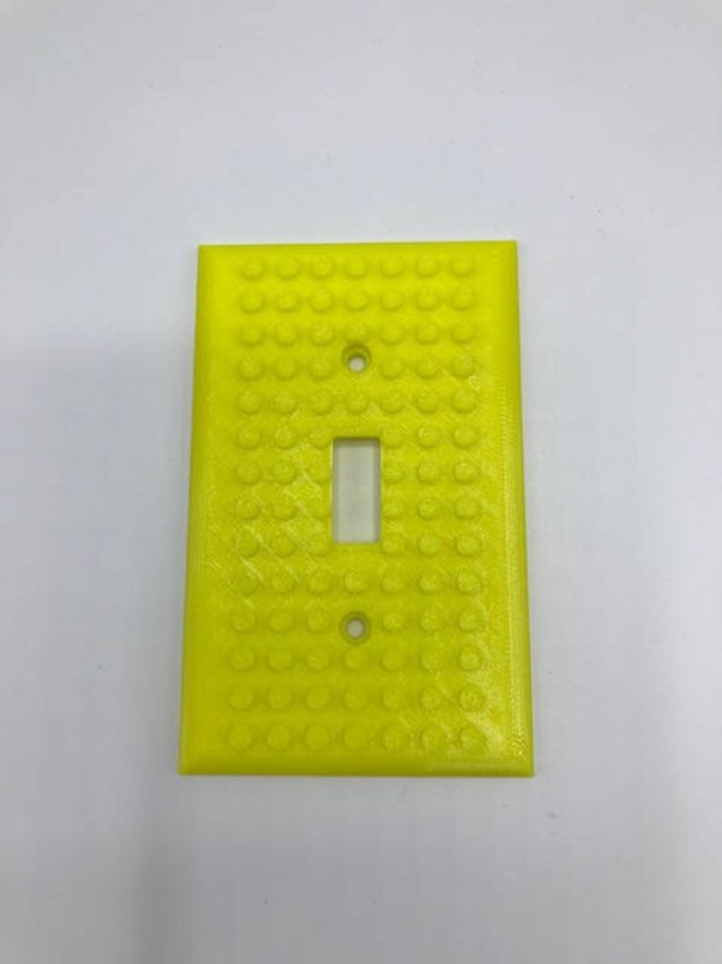 May include: A yellow light switch cover with a textured surface that resembles Lego bricks.