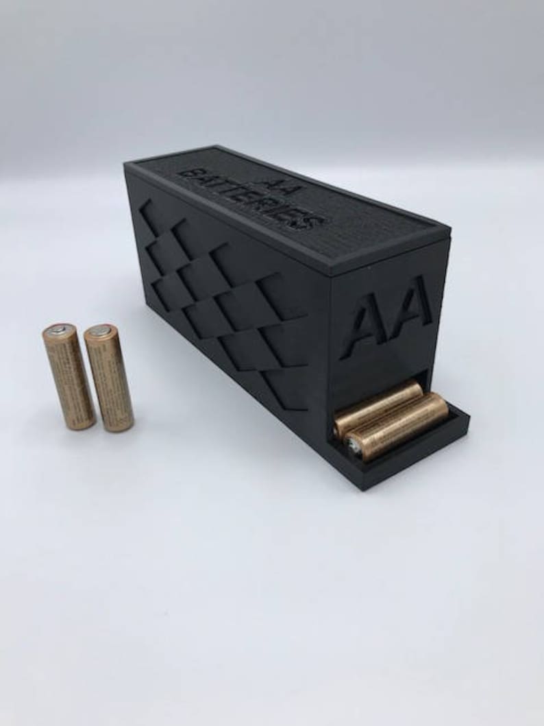 Puede incluir: Una caja de pl&aacute;stico negra con las palabras "AA Batteries" en la parte superior y "AA" en el lateral. La caja tiene un compartimento para guardar las pilas y una ranura para dispensarlas. Hay dos pilas AA doradas sobre una superficie blanca.