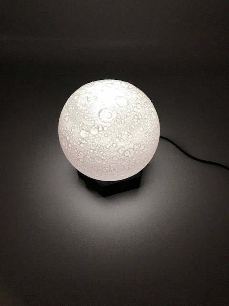 May include: A white moon-shaped lamp with a textured surface, lit up and casting a soft glow. The lamp is on a black base.