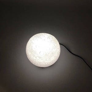 May include: A white moon-shaped lamp with a textured surface. The lamp is lit and casting a soft glow.