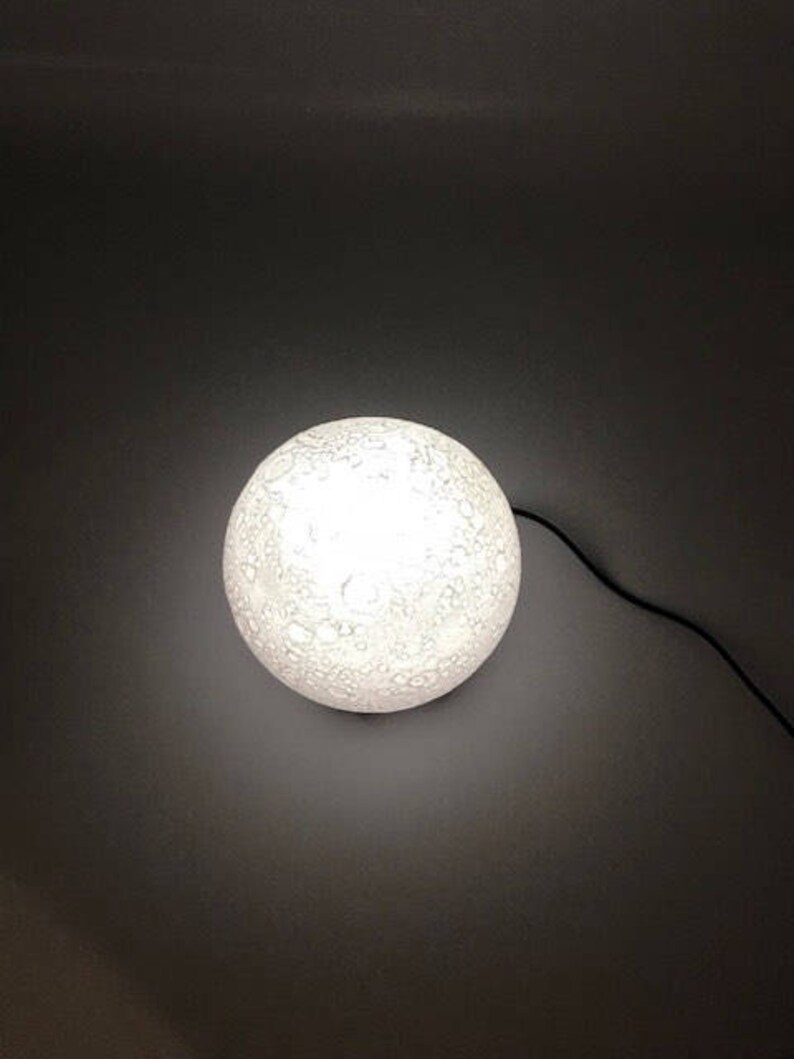 May include: A white moon-shaped lamp with a textured surface. The lamp is lit and casting a soft glow.