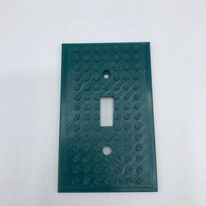 May include: A teal green light switch cover with a textured surface that resembles Lego bricks.