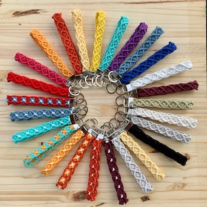 May include: A collection of 20 colorful crocheted keychains with silver key rings. The keychains are arranged in a circle on a wooden surface.