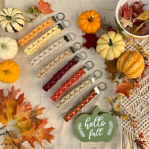 May include: A collection of macrame keychains in various fall colors, including orange, cream, and burgundy, with silver-tone hardware. The keychains are arranged with pumpkins, gourds, and fall leaves, with a wooden sign that says "hello fall".