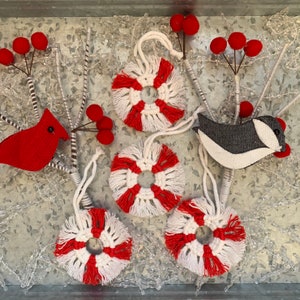 May include: A collection of handmade Christmas ornaments. The ornaments include red cardinals, gray and white birds, and macrame-style circles in red and white. Red berry sprigs are also included.