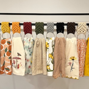 May include: A set of 12 kitchen towels hanging on a black metal rack. The towels are made of different materials and have various patterns, including floral, geometric, and abstract designs. Each towel is attached to a macrame ring.
