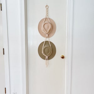 May include: A white macrame hat rack with two hats hanging on it. The top hat is a light beige color and the bottom hat is a light green color. The hat rack is hanging on a white door.