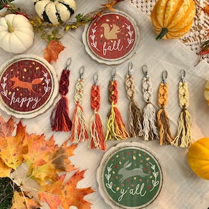 May include: A set of seven colorful macrame keychains with tassels. The keychains are displayed on a table with fall-themed decorations, including wooden signs that say "fall", "happy", and "y'all".