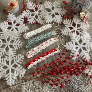 May include: Four crocheted keychains with different color patterns and metal key rings. The keychains are displayed on a surface with white snowflakes and red berries.