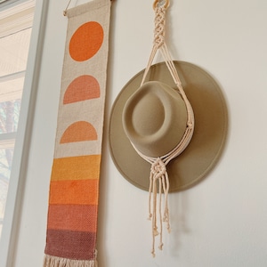 May include: A beige macrame hat hanger with a brown hat hanging from it. A woven wall tapestry with orange, red, and brown geometric shapes is on the wall to the left of the hanger.