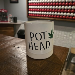 May include: White ceramic mug with the words "POT HEAD" in black, and a green cannabis leaf graphic. The mug sits on a dark wooden surface. The background includes a wall decoration resembling the American flag.