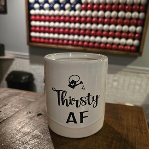 May include: A white ceramic mug with the words "Thirsty AF" in black script and a watering can graphic. The mug is cylindrical and sits on a wooden surface. In the background is a framed American flag made of baseballs.