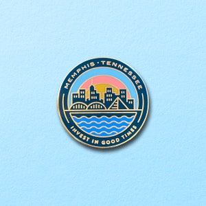 May include: A blue and gold enamel pin with a cityscape silhouette, a bridge, and a sunset. The text "Memphis, Tennessee" and "Invest in Good Times" are written around the pin.