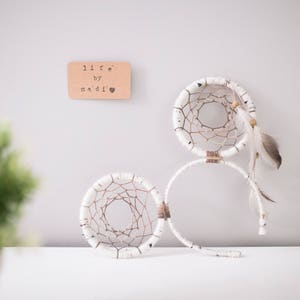 Puede incluir: Dos atrapasueños blancos con centros tejidos y detalles de plumas se exhiben sobre una superficie blanca. Un pequeño letrero rectangular de madera con el texto "life by nadi" está montado en la pared sobre los atrapasueños.
