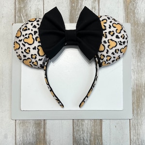 May include: A headband featuring two round ears with a leopard print pattern in tan and black. A large black bow is centered between the ears. The headband itself has a matching leopard print.