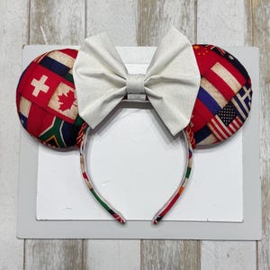 May include: A headband with mouse ears, featuring a large white bow. The ears are covered in a patchwork of international flags, including the USA, Canada, and Switzerland. The headband itself is also decorated with flag patterns.
