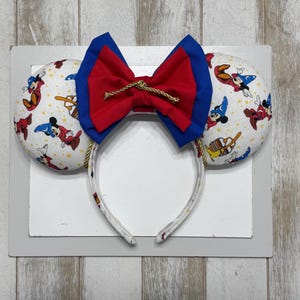 May include: Mickey Mouse ear headband with a red and blue bow. The ears are white with a pattern of Mickey Mouse in various poses. The headband is white with a pattern of Mickey Mouse.