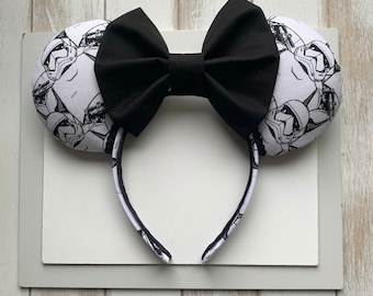 May include: Black and white patterned fabric mouse ears with a black bow. The pattern features a repeated image of a Stormtrooper helmet.