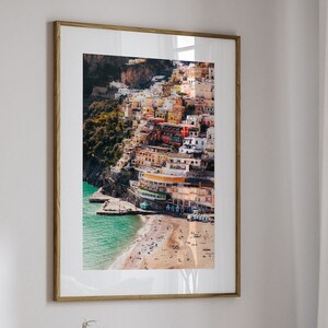 May include: A framed photograph of a colorful Italian village built on a cliffside overlooking a beach. The photo is hanging on a white wall in a living room setting with a white couch, a throw blanket, and a coffee table.