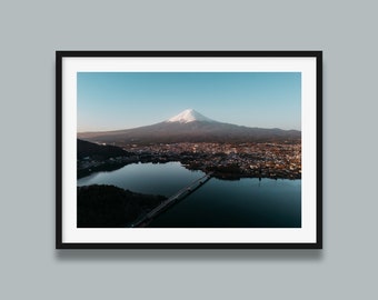 Può includere: Una vista aerea del Monte Fuji, un vulcano innevato in Giappone, con un ponte e un specchio d'acqua in primo piano.
