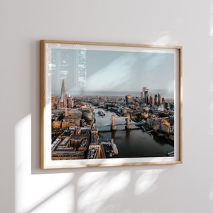 May include: An aerial view of the London skyline featuring the Tower Bridge and the Shard in the distance. The River Thames flows through the city, with numerous buildings and bridges visible.
