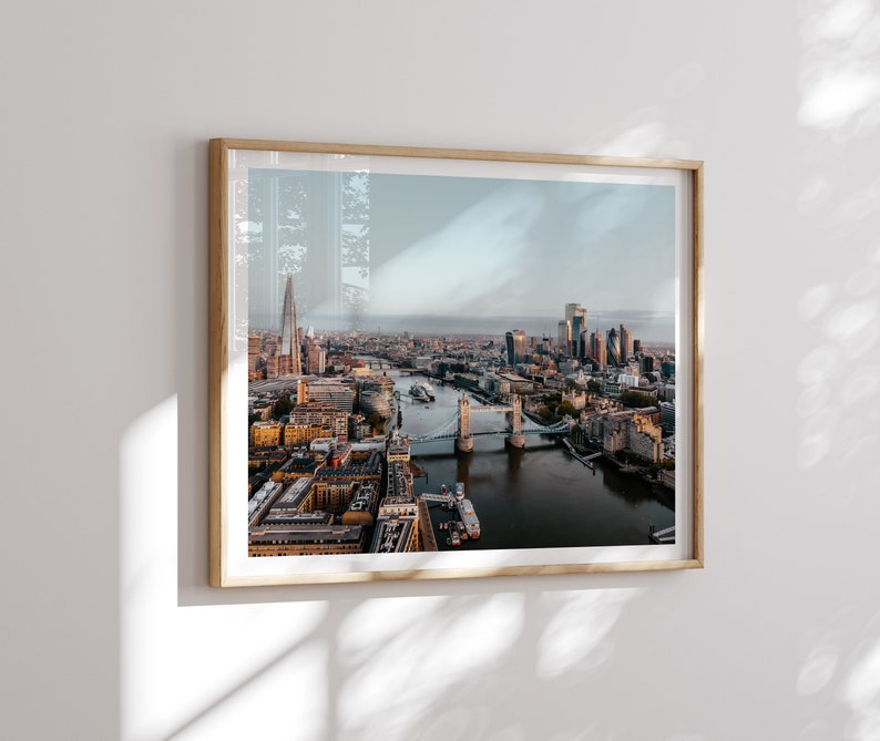 May include: An aerial view of the London skyline featuring the Tower Bridge and the Shard in the distance. The River Thames flows through the city, with numerous buildings and bridges visible.