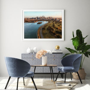 May include: A framed aerial photograph of a city skyline with a curved road leading to a green park. The photo is hanging on a white wall above a gray dresser with two blue chairs and a wooden coffee table.