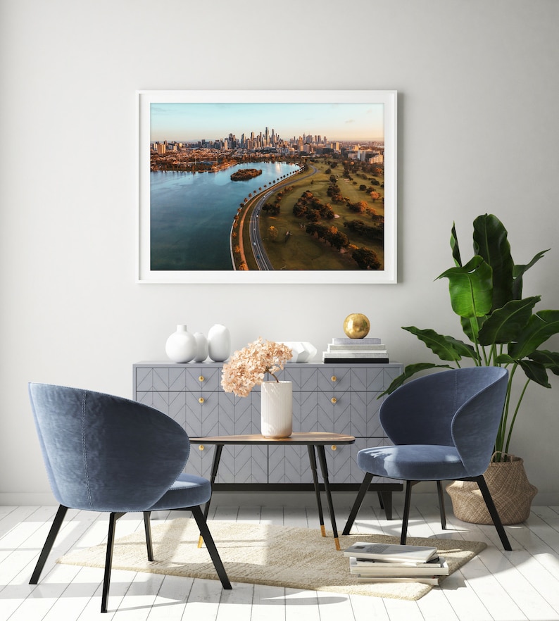 May include: A framed aerial photograph of a city skyline with a curved road leading to a green park. The photo is hanging on a white wall above a gray dresser with two blue chairs and a wooden coffee table.