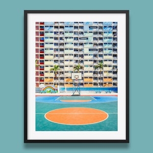 Op de afbeelding: Een kleurrijk appartementencomplex in Hongkong met een basketbalveld ervoor. Het veld is geschilderd in tinten blauw, groen en oranje. Er is een basketbalring in het midden van het veld.