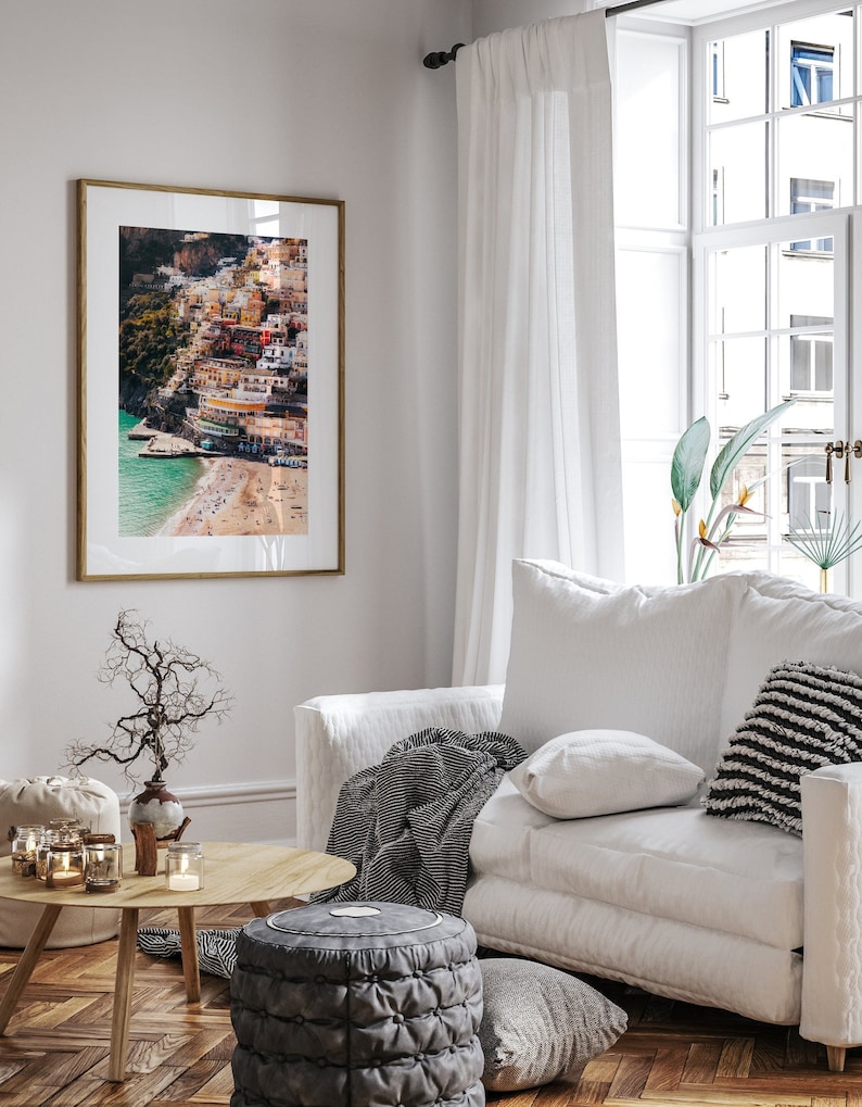 May include: A framed photograph of a colorful Italian village built on a cliffside overlooking a beach. The photo is hanging on a white wall in a living room setting with a white couch, a throw blanket, and a coffee table.