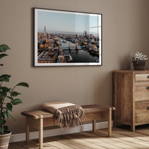 May include: A framed photograph of the London skyline with Tower Bridge in the foreground. The photo is hanging on a beige wall above a wooden bench with a brown throw blanket.