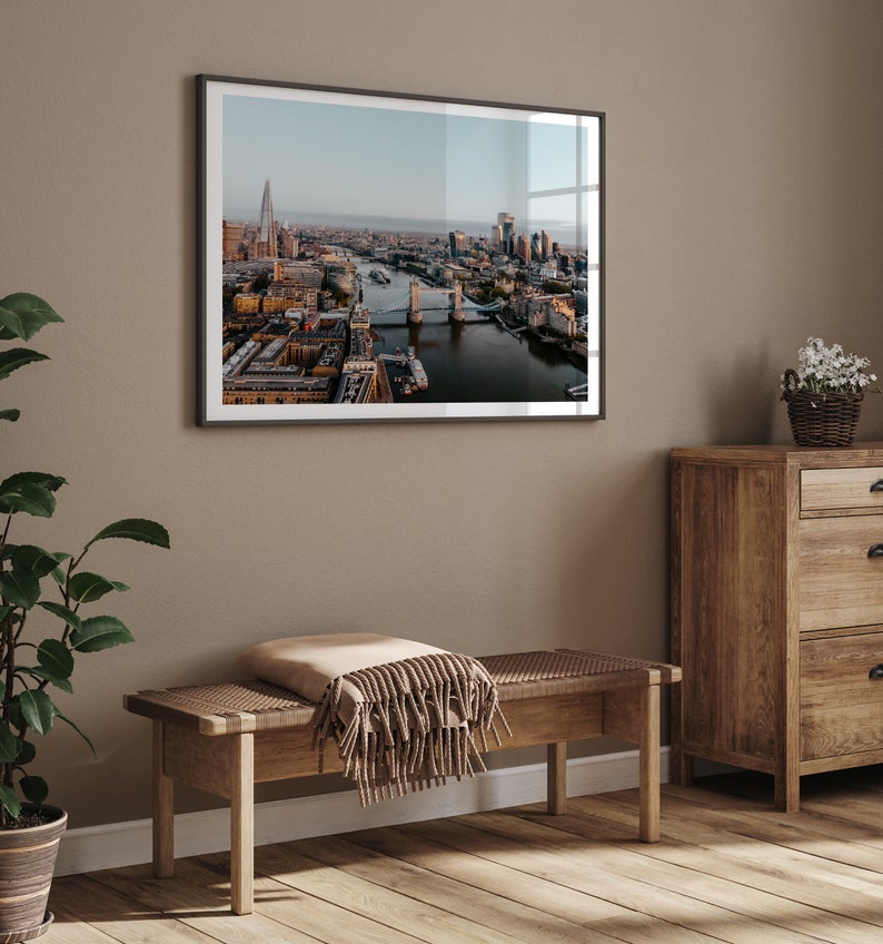May include: A framed photograph of the London skyline with Tower Bridge in the foreground. The photo is hanging on a beige wall above a wooden bench with a brown throw blanket.