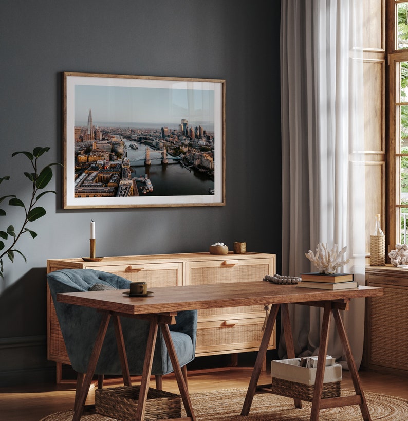 May include: A framed photograph of an aerial view of London, England, showing Tower Bridge and the River Thames. The photo is hanging on a dark grey wall above a wooden dresser and a blue armchair.