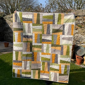 May include: A handmade quilt with a patchwork design. The quilt features a variety of fabrics in shades of green, gray, and mustard yellow. The quilt is rectangular and appears to be approximately 70 inches by 70 inches.