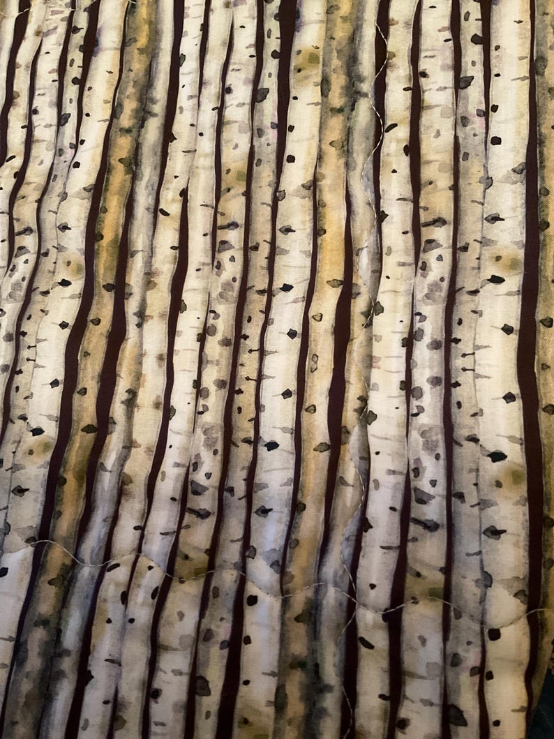 K&ouml;nnte beinhalten: Ein gesteppter Stoff mit einem Birkenbaum-Design. Das Muster zeigt vertikale Linien, die Baumst&auml;mmen in Braun-, Beige- und Schwarzt&ouml;nen &auml;hneln. Der Stoff hat ein strukturiertes Aussehen, mit einem sichtbaren Steppmuster.