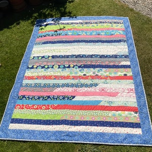 Op de afbeelding: Een kleurrijk patchwork-deken met een blauwe rand en verschillende bloemen- en geometrische patronen. Het deken is gemaakt van stroken stof die in een horizontaal patroon aan elkaar zijn genaaid.