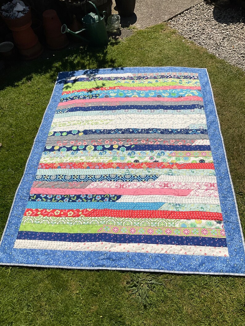 Op de afbeelding: Een kleurrijk patchwork-deken met een blauwe rand en verschillende bloemen- en geometrische patronen. Het deken is gemaakt van stroken stof die in een horizontaal patroon aan elkaar zijn genaaid.